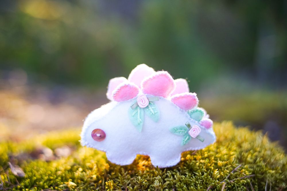 A white stegosaurus with pink plates and roses on its body. The stegosaurus has pink eyes.
It's standing on green moss.