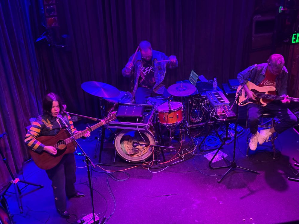 A live music performance featuring three musicians on stage. One musician plays an acoustic guitar and sings, while the other two are positioned behind drums and an electric guitar, respectively. The stage is illuminated with purple lighting, and various instruments and equipment are visible.