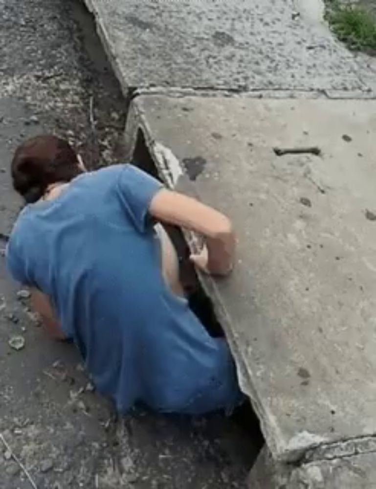 a woman in a blue shirt is crawling out of a hole in a sidewalk .