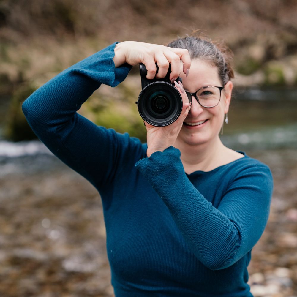 Mitte 30 Jährige Fotografin mit türkisfarbenem Pulli hält eine Kamera richtung Betrachter und lächelt. Im Hintergrund ist verschwommen ein Bach und ein Wald zu sehen, die Fotografin steht auf einer Kiesbank. man sieht aber nur ihren Oberkörper 