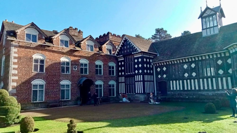 Photograph of Rufford Old Hall in Lancashire. Distinct C16th Tudor Great Hall wing in its black and white finery adjoined by its newer C18th accommodation block of soft red brick symmetrical features. In front is a green lawn. Blue sky overhead.

On the far right of the photograph appears a dinosaur. It isn’t but a person in a padded coat taking a photo 😂