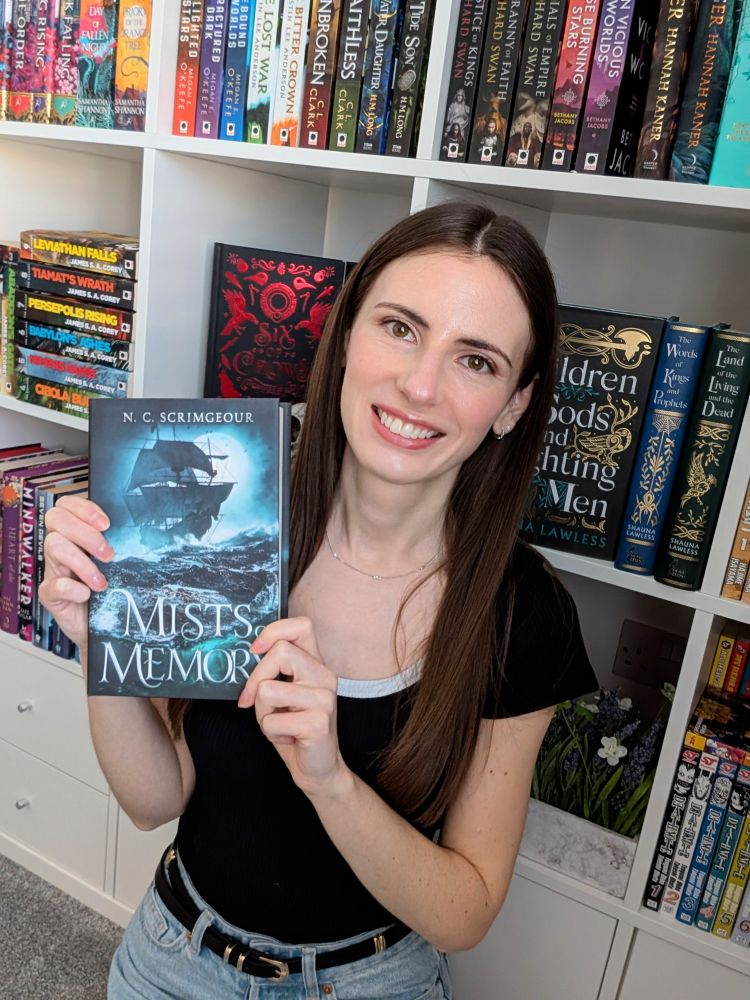 N. C. Scrimgeour in front of a bookshelf, holding a copy of her new book, Mists of Memory 