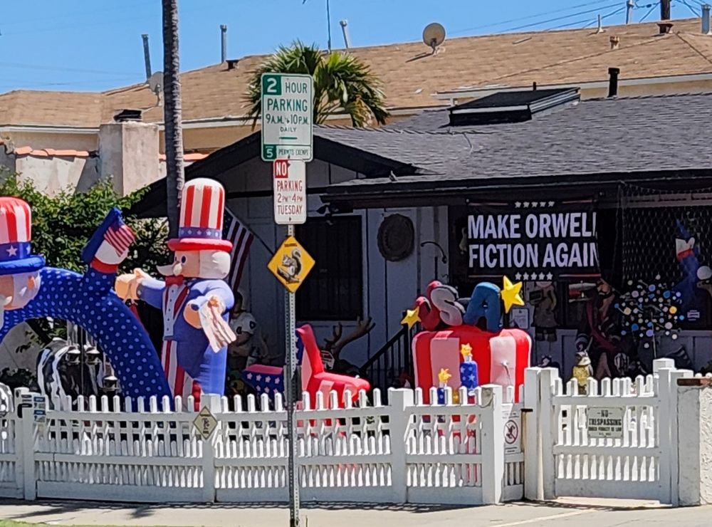 A front yard with air filled inflatable uncle Sam and other American flag colored inflatables. Banner says Make Orwell Fiction Again.