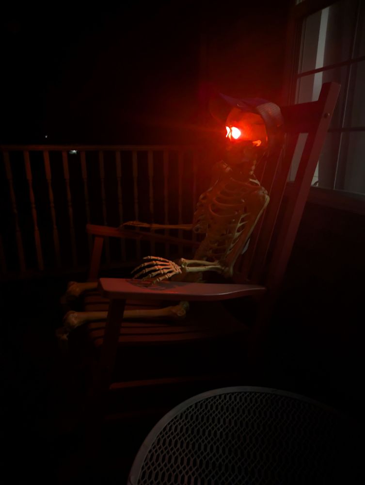 A photo of a skeleton, wearing a Detroit Tigers ball cap, sitting in a rocking chair on the front porch. Part of a round blue table is visible. The skeleton has glowing red eyes. The photo was taken in the dark and it looks eerie.