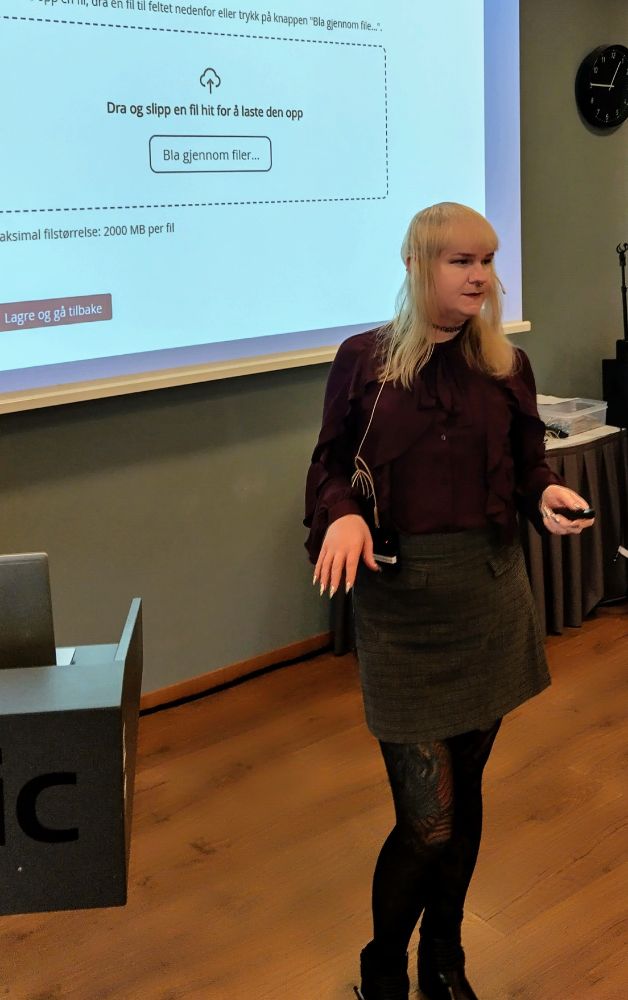 white woman with platinum blonde hair with bangs holding a presentation in front of a screen while wearing a burgundy blouse with frills, a short tweed skirt and black pantyhose with her leg tattoos visible through it.