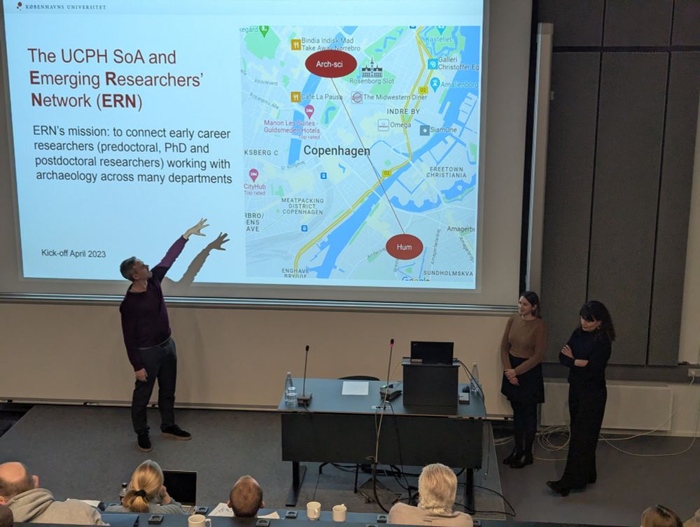 A presentation slide showing information about UCPH SoA and Emerging Researchers' Network (ERN), with a map of Copenhagen marking different department locations. A presenter gestures enthusiastically at the map while explaining ERN's mission to connect early career researchers across departments. Two other presenters stand to the side of the podium.