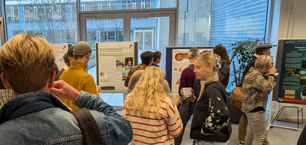 A crowded poster session during a conference break, showing attendees examining various research posters along a windowed corridor. Several posters are visible, including one titled 'Research of the Human Remains' and 'The Hidden Pot'. Students and researchers, holding coffee cups, are engaged in viewing the displays.