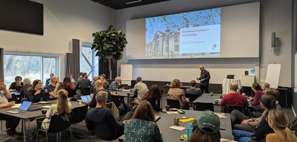 Workshop at the School of Archaeology taking place on March 25. The image shows a modern classroom with attendees seated at tables with laptops and notebooks. A presenter, likely Christian Tangkjær, stands at the front addressing the group. A projection screen displays the workshop title and date. The room has large windows overlooking trees, and various presentation materials including flip charts are visible. The workshop is focused on discussing lifelong learning initiatives as part of the University of Copenhagen's 2030 strategy.