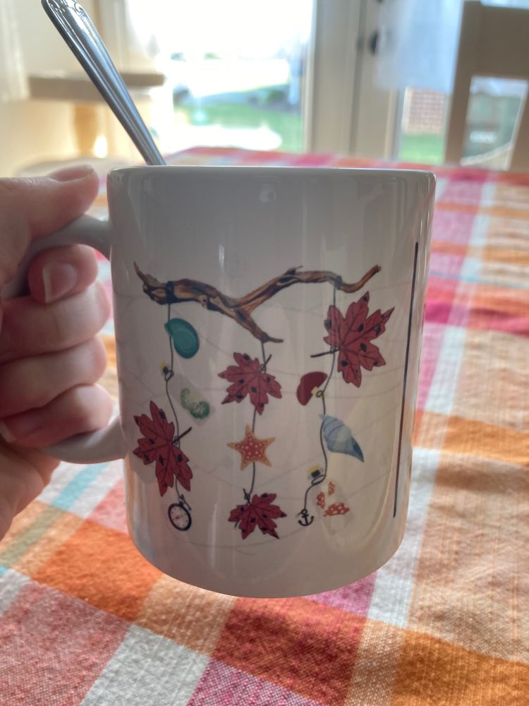 A hand holding a mug with a design of maple leaves and seashells. The leaves have spots on them. 