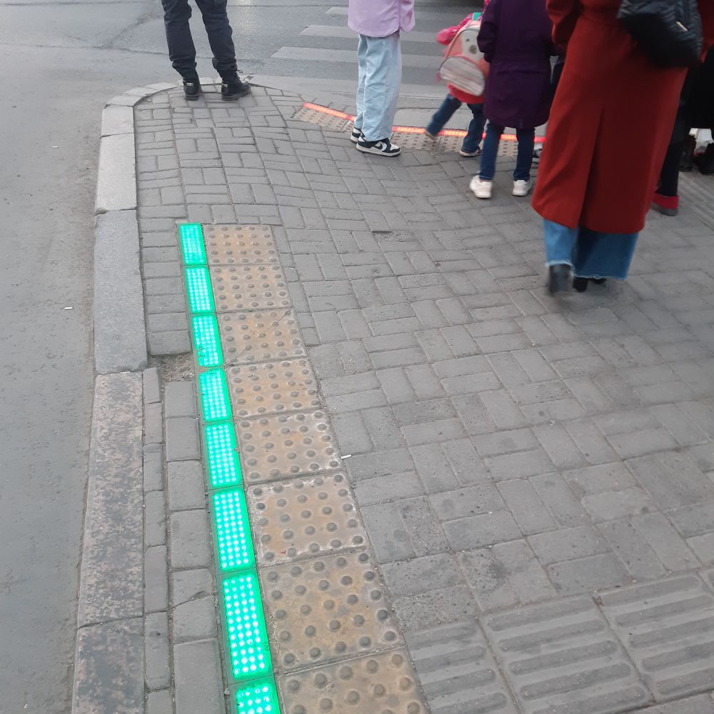 8 25cm or so long, 5cm wide strips of LEDs, currently light in green, though you can see red set in the background. Set in sidewalk with tactile tiles, bumpity for edge of road, lines for safe walking toward crossing. 