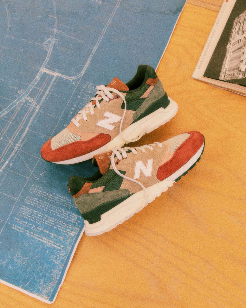 New Balance 998s in an earthy tone with hits of orange and green, shot in a wood table with a blueprint 