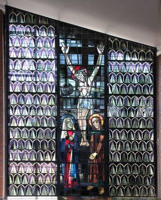 A stained-glass triptych showing Christ on the cross in the center; to left and right geometric patterns in blue and green. Photo via Forschungsstelle Glasmalerei des 20. Jahrhunderts, https://www.glasmalerei-ev-web.de/pages/b6652/b6652.shtml