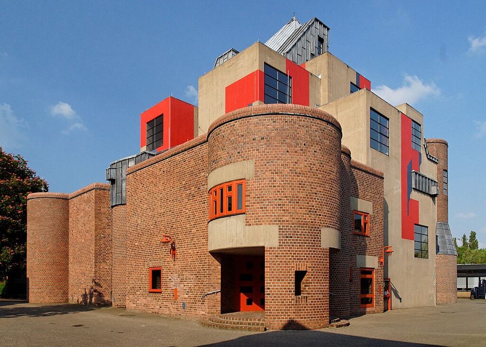 A brick and concrete church. The main mass is of CIP concrete with swathes of the exterior painted red. The entrance is at the round corner tower of a brick wing. The structure is irregular and varied. Photo by Wiegels via Wikimedia Commons.  
