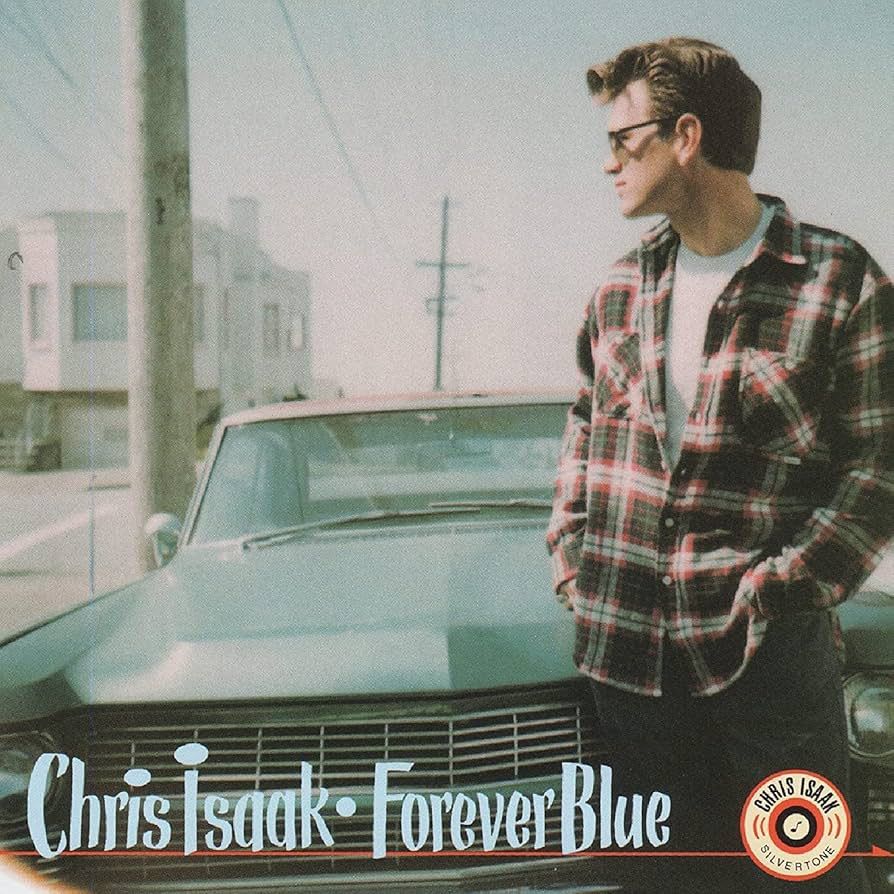 chris isaak wearing jeans, white t shirt, flannel shirt and shades. He’s leaning on the hood of a Cadillac (?) as the fog rolls in in the Sunset in San Francisco 