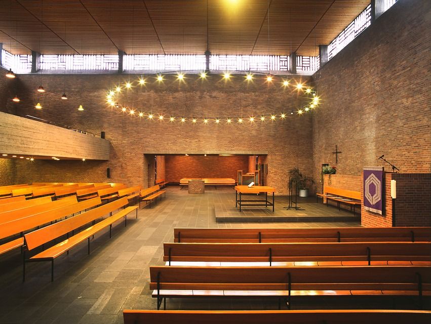 A worship space. To the right is a, altar raised by one step. Immediately in front of the photographer and to the left are wooden benches. There is a narrow band of clerestory windows and a wooden ceiling. In the back of the photo, on the other side of a baptismal font, is a smaller space with benches. A ring chandelier hangs above the center of the space. Photo by LVR-Amt für Denkmalpflege im Rheinland, Jürgen Gregori via Straße der Moderne, https://strasse-der-moderne.de/kirchen/krefeld-bockum-christuskirche/
