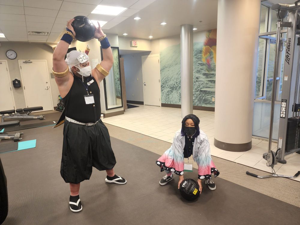 Tengen Uzui and Shinobu Kocho cosplayers in a hotel fitness center. Tengen is holding a medicine ball over his head, while Shinobu struggles to lift one off the floor.