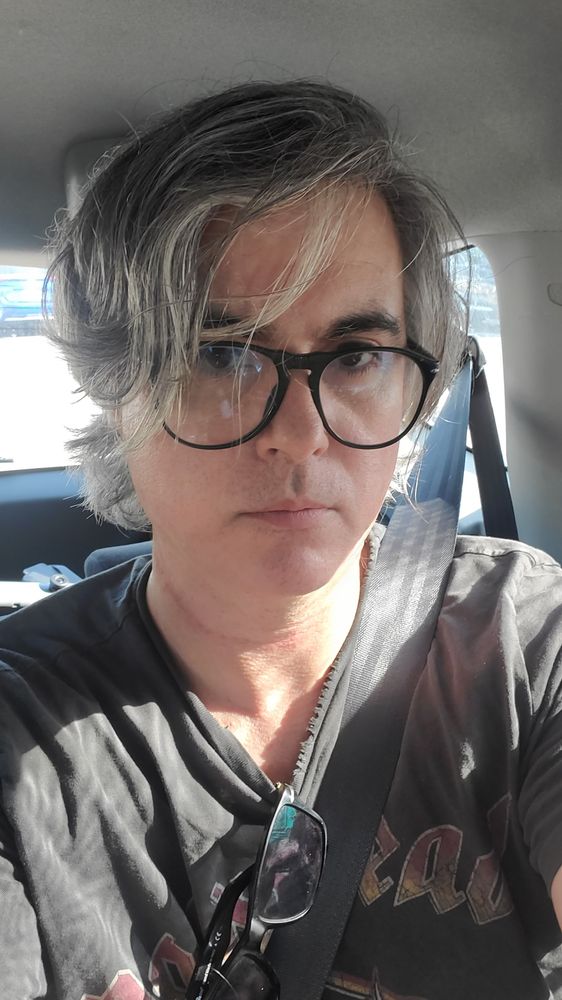 A selfie of me sitting in the back of a car with my seat belt on. Black shirt with sunglasses hanging off them.