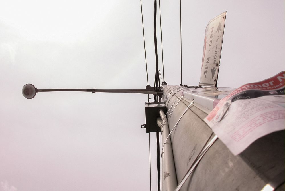 Upward view of a streetlight pole with electrical wires crossing horizontally against a pale, overcast sky.