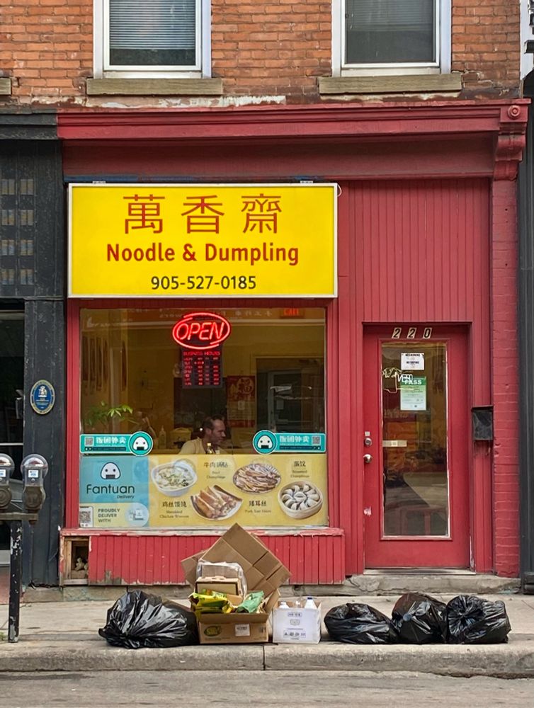 Red shopfront with yellow sign, promising noodle and dumpling, singular. The bottom half of the display window shows a number of dishes and their prices. Above this menu a man can be seen eating at a window seat.