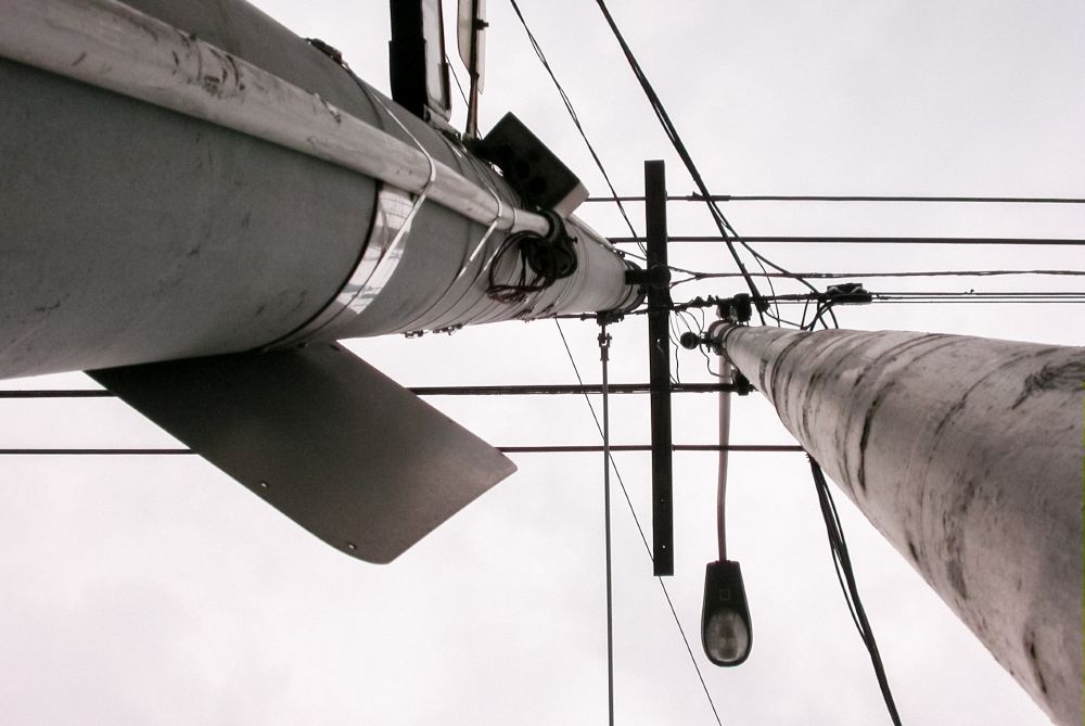 Upward view of a streetlight pole with electrical wires crossing horizontally against a pale, overcast sky.