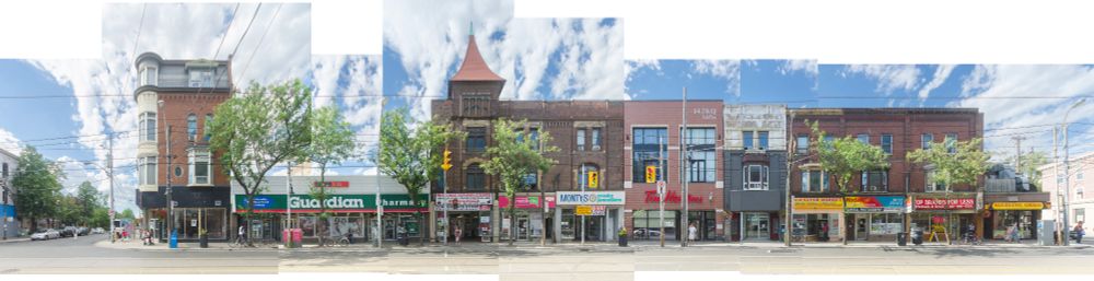 Stitched panoramic image of a city street lined with brick buildings. Storefronts include a pharmacy, coffee shop, and various retail outlets. Trees line the sidewalk under a bright, cloudy sky.
