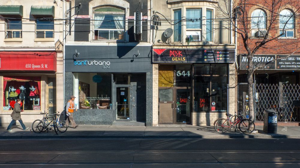 Street view of a row of buildings with ground floor storefronts. Signs for a spa and a shop are visible. People walk by, and two bikes are parked nearby. Sunny day.