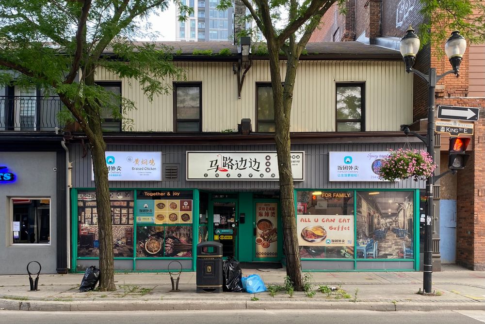 Street view of a two-story building with signage in English and Chinese, Offering braised chicken and an "All U Can Eat Buffet." Trees and a streetlamp decorate the scene.