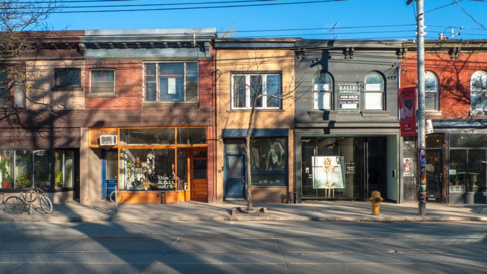 Row of colorful brick storefronts on a sunny street, featuring a crocheted cafe, a "For Sale" sign, and a bicycle against a window display. 