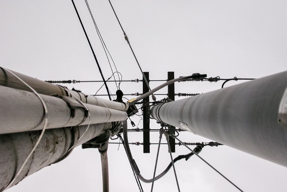 Upward view of two tall utility poles with interconnected power lines against a cloudy sky. One appears to be reaching towards the other.
