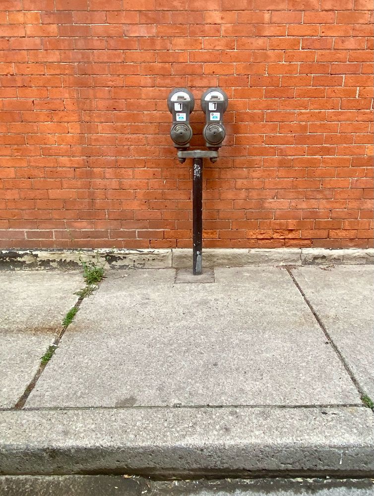 Two parking meters sharing a single post are installed on a sidewalk but set back from the curb, close to a brick wall.