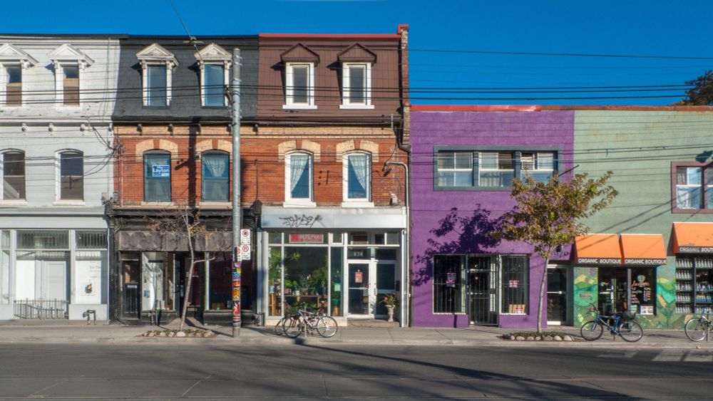 Colourful urban street view with old brick buildings. A row of three-story Victorian buildings with third-floor dormers ends at a two-story flat-roofed building. Purple and green facades house shops; bicycles parked out front. Clear blue sky. Vibrant.