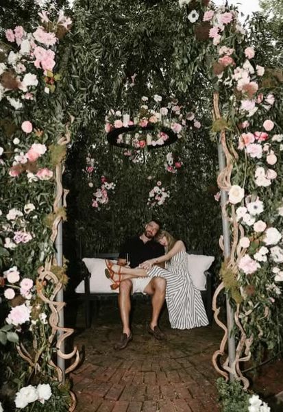 Taylor and Travis sitting in a floral archway, she’s resting her legs over his