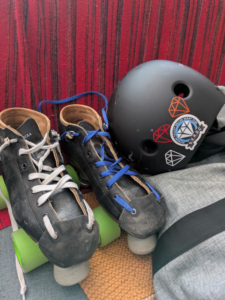A pair of well-lobed and worn black roller derby quad skates with bright green wheels. The left boot is laced up with white laces, the right boot laces up with blue laces. Next to the skates is a black helmet with 3 diamond stickers on it; one white, one red and one orange, each sticker represents passing a skill level. The coloured diamond stickers surround a bigger diamond sticker with a scroll bearing club name: Diamond Valley Roller Derby Club. 