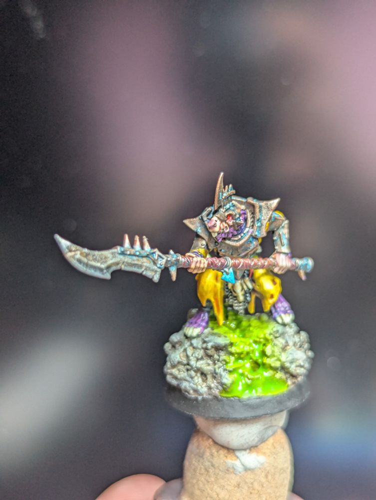 A photo of a Skaven miniature from Age of Sigmar. It is a little rat creature in armor holding a spear. The armor is sort of a copper color but worn with verdigris on it. The spear is rusted. He is standing on a rocky base offer a vibrant yellow green slime