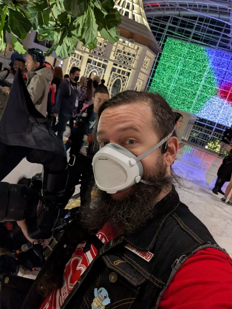Scrane, a white, bearded,asc presenting person with long brown hair and a long brown beard poses in front of a white gazebo indoors while holding his camera