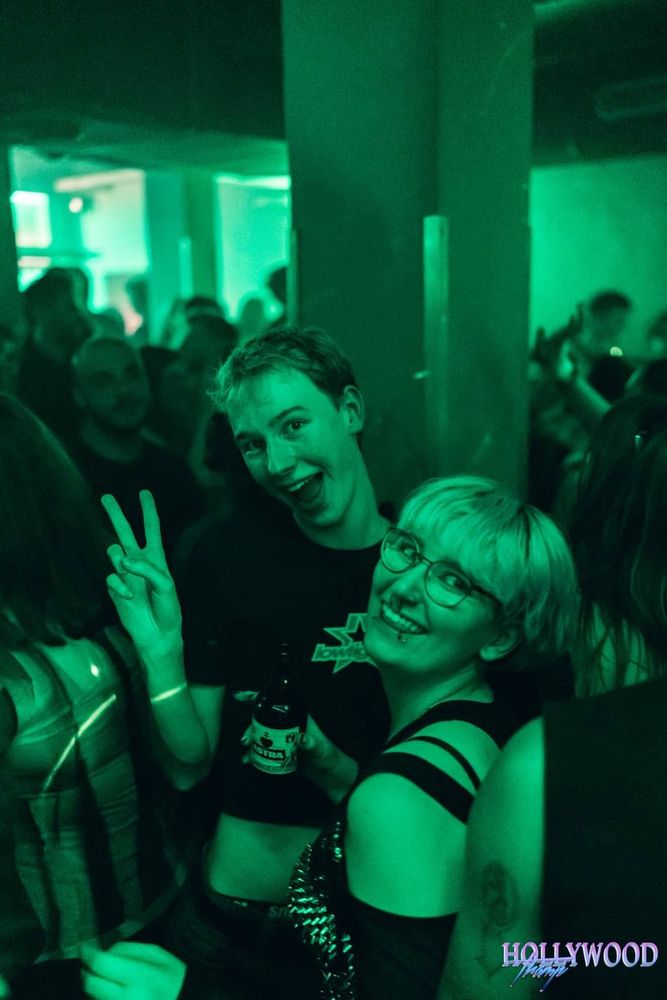 A green lit club ambiance with two people in the foreground. One is throwing up a peace sign while the other is grinning at the camera with a beer in their hand, both are smiling widely. 