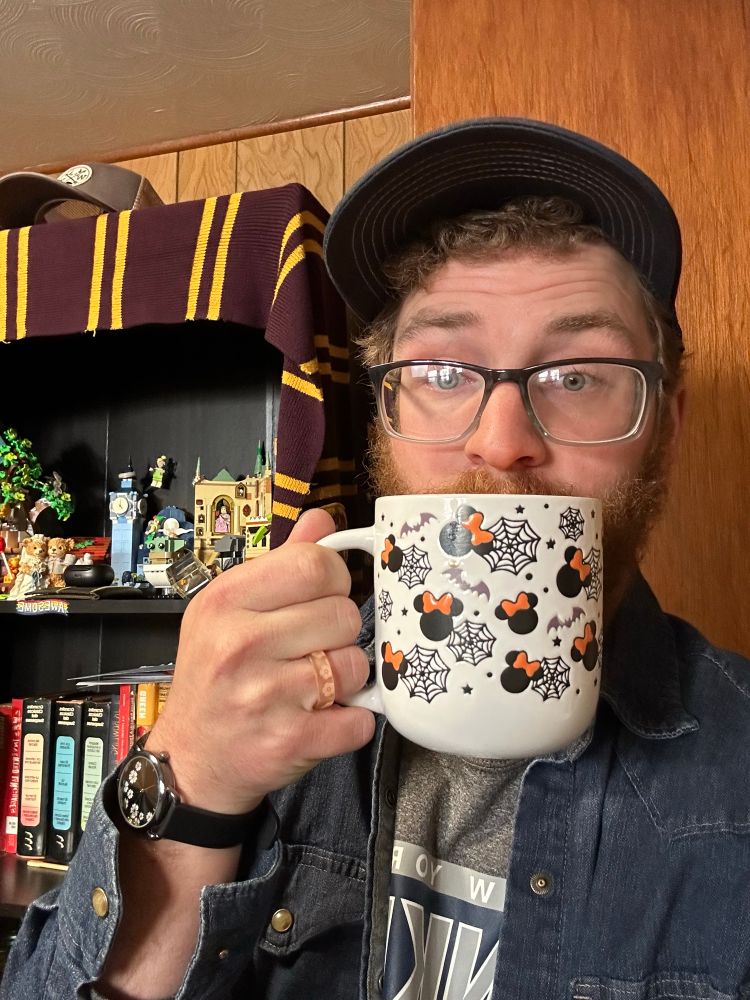 Louis, wearing a denim button up over a Yankees shirt, and a baseball cap. Holding a coffee mug patterned with spider webs, bats, and Minnie Mouse heads. Standing in front of a full bookcase. 