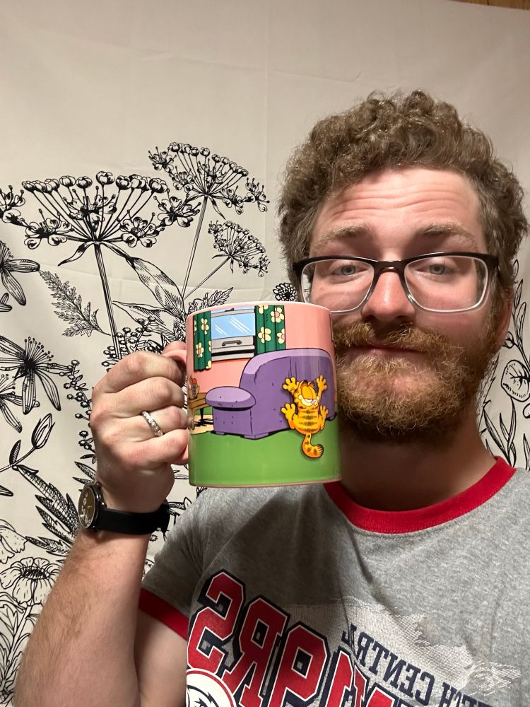 I’m standing in front of a black and white floral tapestry holding a mug depicting Garfield the cat. I’m wearing a grey short sleeve shirt with red accents. On my wrist is a black watch. 