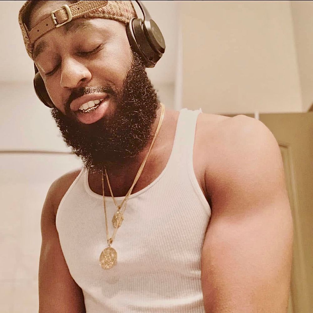 A baddie with a stylish beard, headphones on, and a shiny gold chain known as Zaddy is immersed in his music. He is showcasing muscular arms, a gold sexy mouth grill and a polo hat backwards. His eyes are closed, yet he sees it all. You! 