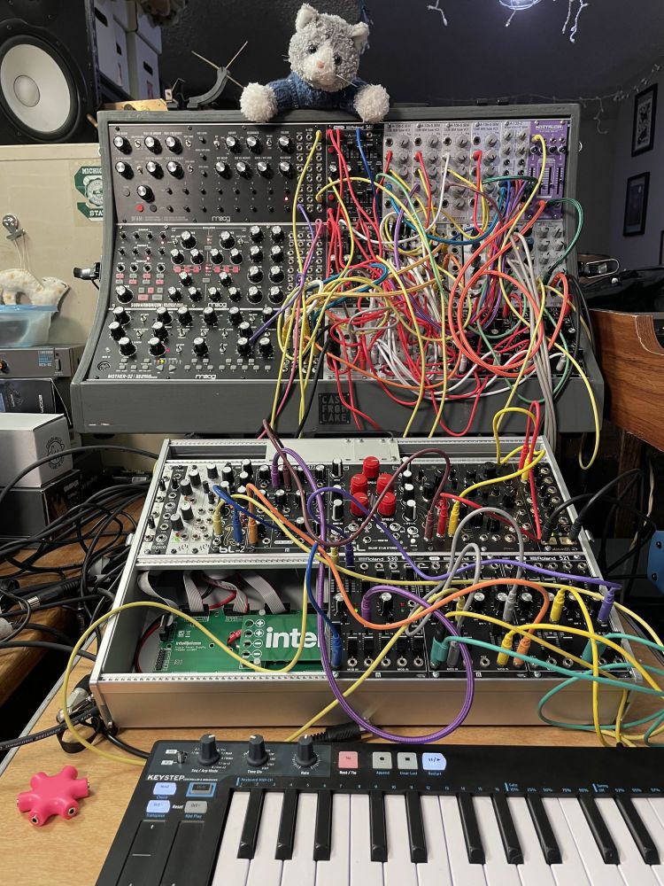 Two Eurorack-format racks with a mess of colorful cables and a small plush cat perched on top.