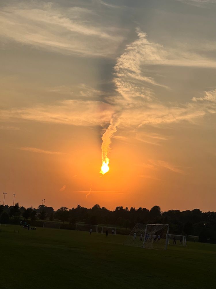 Picture of sunset. The sun is behind a jet trail and it looks like the sun is giving off smoke. 