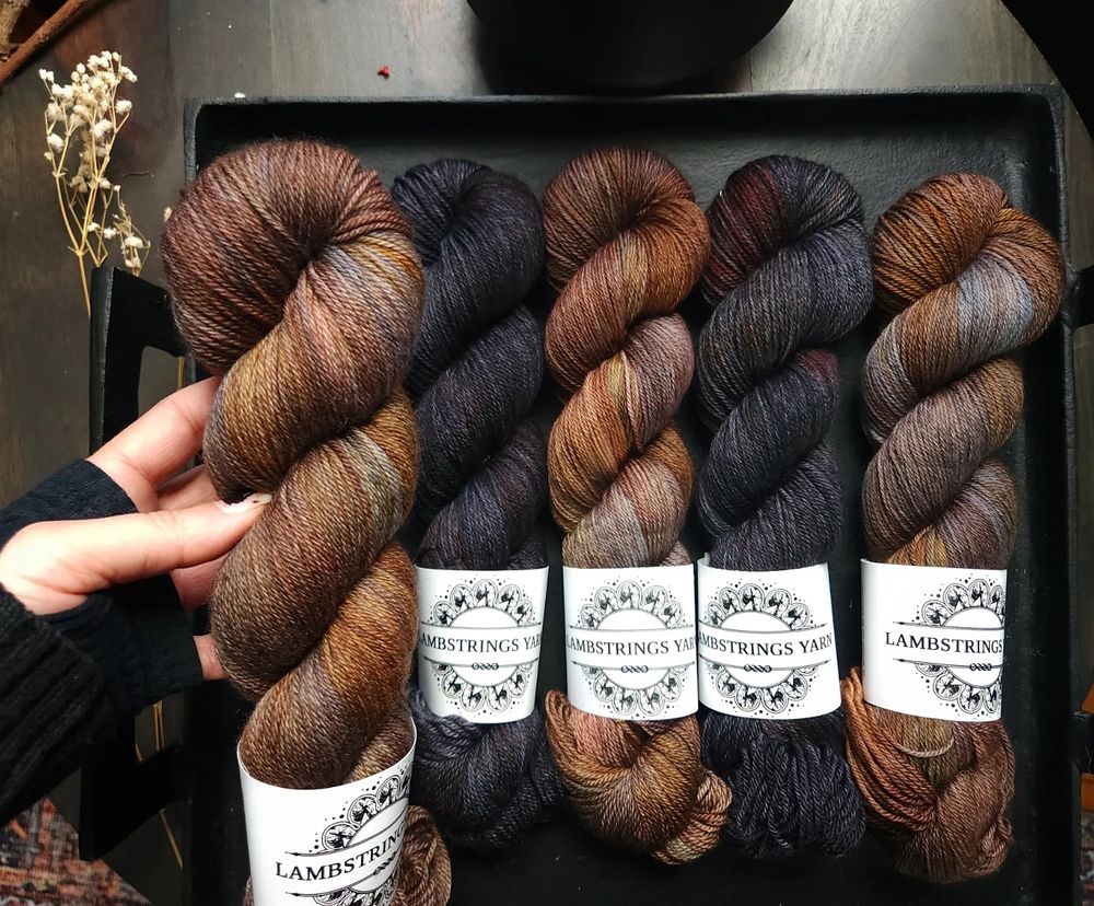 A white woman’s hand holding an earthy skein of yarn in brown tonals, below are alternating skeins in the same color and a deep slate color.