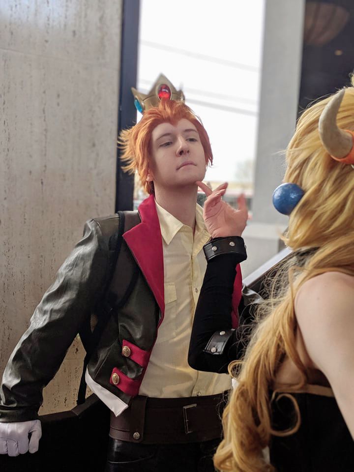Red haired Prince in a green and red, reptilian jacket and a yellow shirt and a long blonde haired princess with Bowser horns and spiked cuffs lifting their prince’s chin with their finger.  