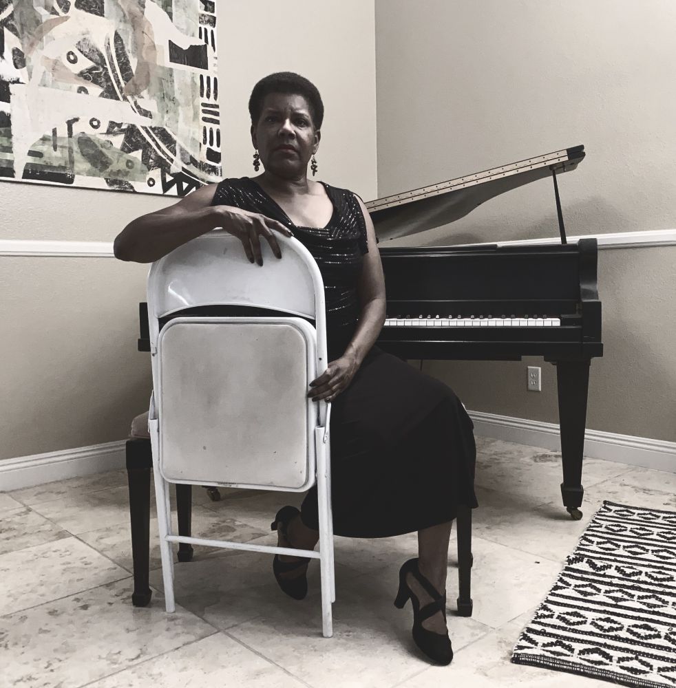 Tananarive posing with a white folding chair in front of her piano. 