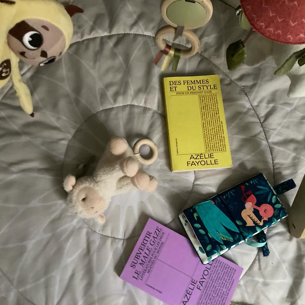 sur le tapis d'activité du bay, quelques éléments de son arche sur le cadre supérieur, un joujou, des anneaux en bois et des peluches, un petit mouton musical et deux livres, Des femmes et du style et le prochain Subvertir le male gaze, à couverture lavande, entrecroisés avec un mini livre en tissu
