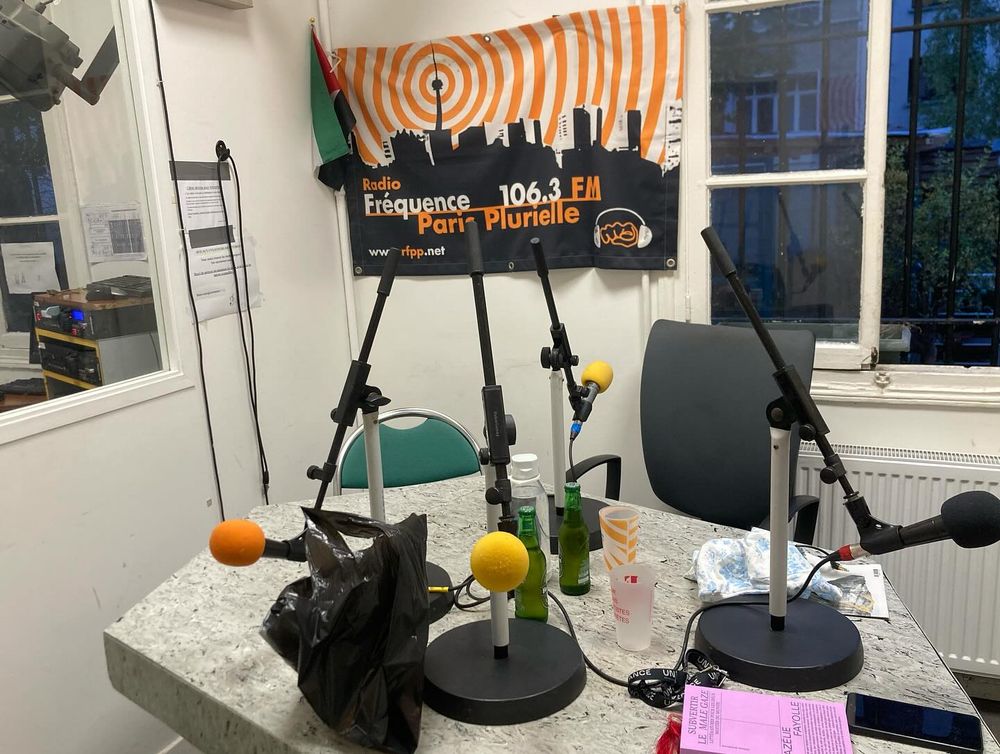 Photo du studio de FPP, avec une bannière en tissu à silhouette urbaine noire et ondes oranges, des micros de radio, des gobelets, deux bières et Subvertir le male gaze sur la table