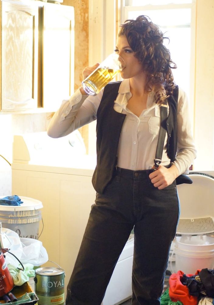 Sun-soaked photo of me, a tall white lady with voluminous curly hair and fake eyelashes, wearing black pants, suspenders and a white dress shirt and drinking beer out of a tall glass. I'm standing in a storage room filled with a cabinet, random buckets of paint and, inexplicably, a (laundry) washing machine