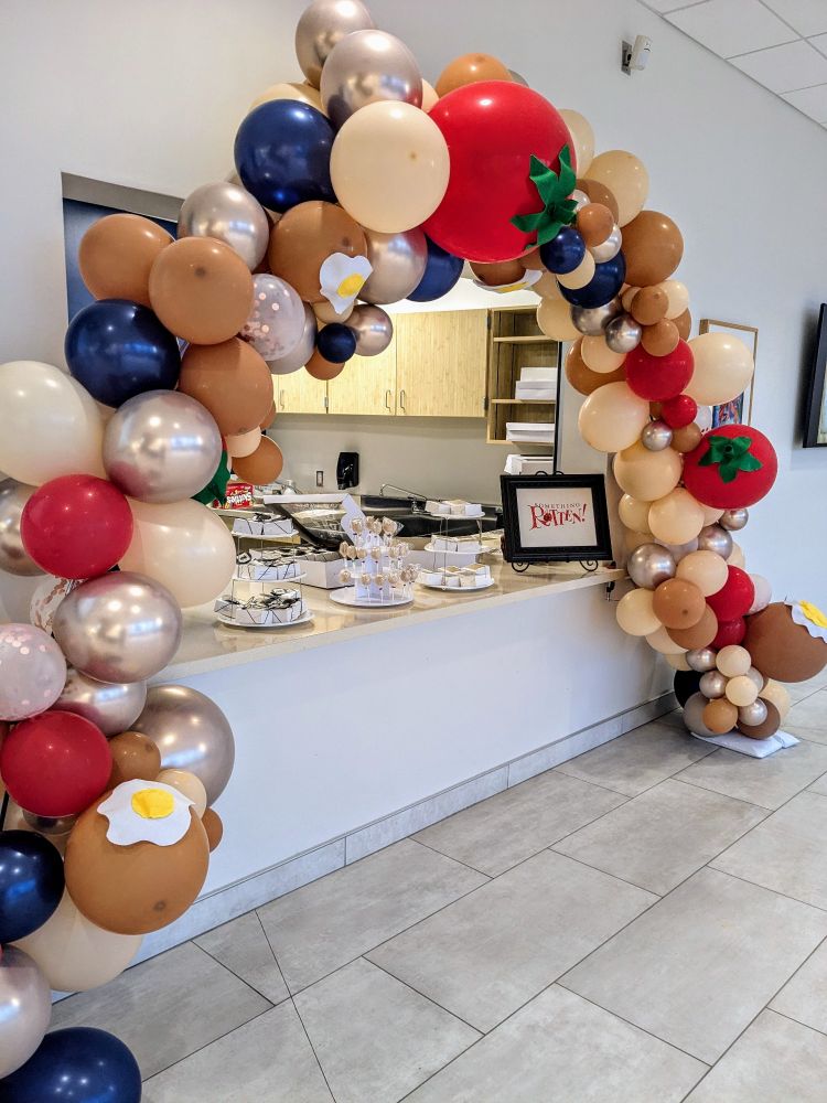 Balloon arch over a small indoor concession stand. The red balloons have green felt stems added so that they look like tomatoes, and there are felt sunny-side up eggs on some of the beige balloons.