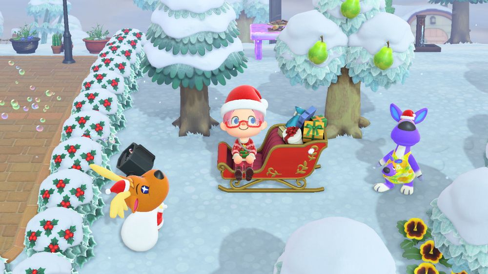 Foto de Animal Crossing New Horizons en la que se ve a mi personaje sonriente sentado en un trineo cargado de regalos, llevando un gorro de Papá Noel, gafas rojas y un jersey navideño rojo con un árbol de navidad dibujado. Todo a su alrededor está nevado, viéndose una hilera de arbustos de acebo a la izquierda rodeando la plaza de la isla. A la izquierda de la imagen, junto al acebo, se puede ver al simpático reno Renato con un gorrito de Papá Noel y su saco blanco cargado de regalos a la espalda. A la derecha de la imagen está Cándida, una canguro de color violeta que también lleva un gorrito de Papá Noel.