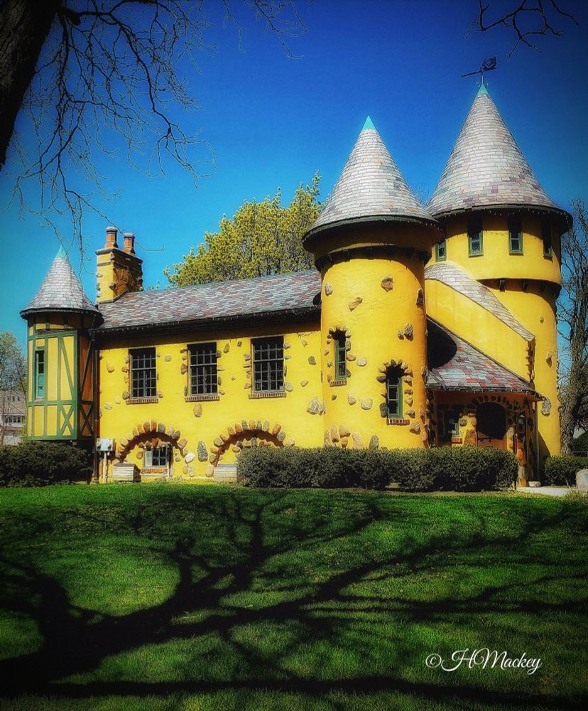 A small castle with a yellow stucco exterior with randomly placed decorative fieldstones. There are spires at three corners, and it has a slate-covered gable roof with a chimney. Some shrubbery hugs the corners the building. A green lawn with the shadow of outstretched branches from a large tree dominates the foreground.